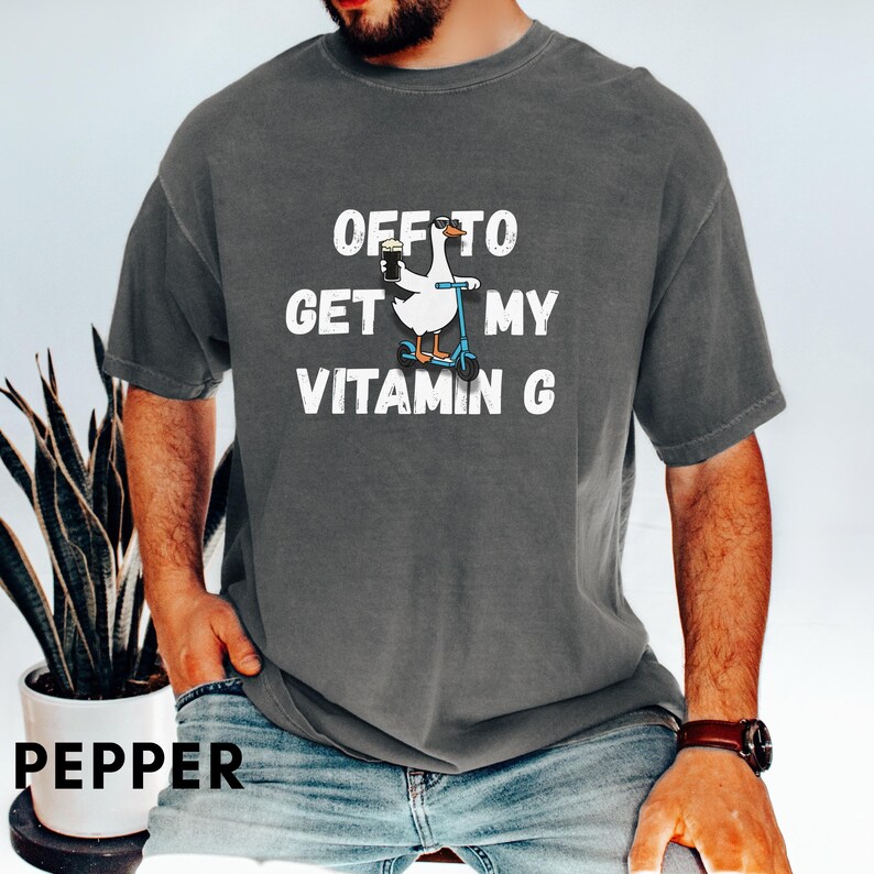 Puede incluir: Camiseta color pimienta con el texto "OFF TO GET MY VITAMIN G". El dise&ntilde;o presenta un ganso blanco montado en un scooter y sosteniendo una bebida. Es una prenda de vestir informal.