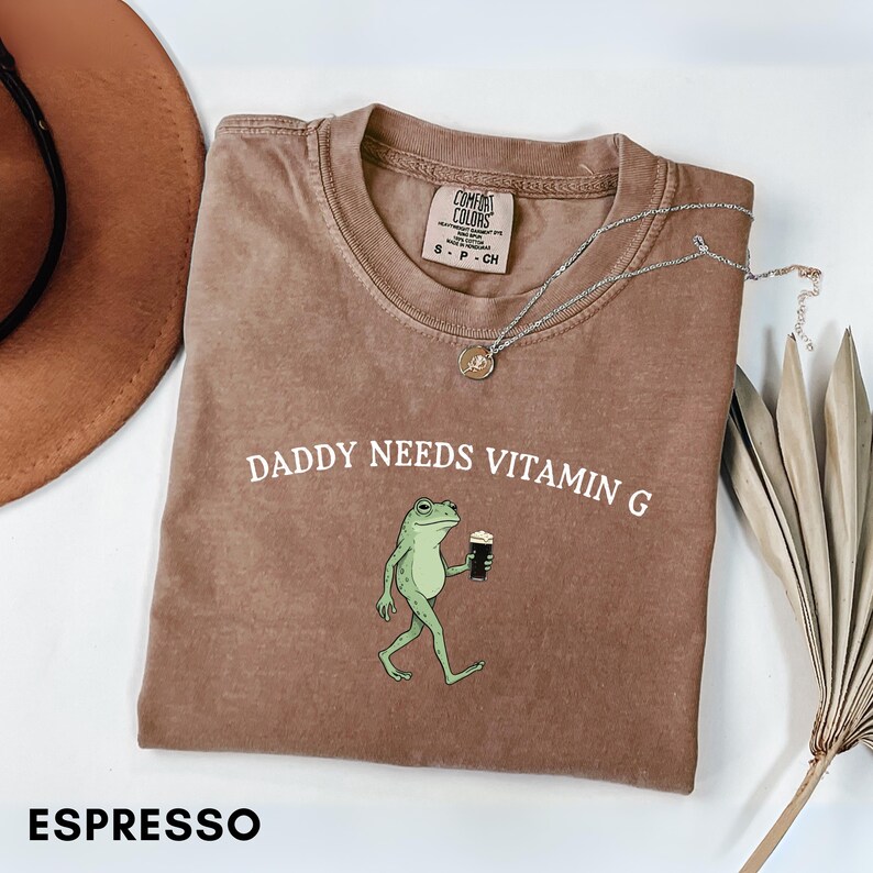 K&ouml;nnte beinhalten: Espressofarbenes T-Shirt mit dem Text "DADDY NEEDS VITAMIN G" und einem Cartoon-Frosch, der ein dunkles Getr&auml;nk h&auml;lt. Das Shirt hat einen Rundhalsausschnitt und ein Comfort Colors-Etikett.