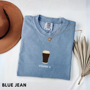 Pu&ograve; includere: Una maglietta blu jeans Comfort Colors con una grafica di una birra scura in un bicchiere e la scritta "VITAMIN G". Una collana d'oro e un cappello marrone sono anche nell'immagine.