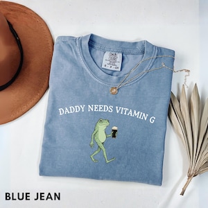 K&ouml;nnte beinhalten: Blaues Jeans Comfort Colors T-Shirt mit dem Text "DADDY NEEDS VITAMIN G" und einem Cartoon-Frosch, der ein dunkles Getr&auml;nk h&auml;lt. Eine goldene Halskette und ein brauner Hut sind ebenfalls sichtbar.