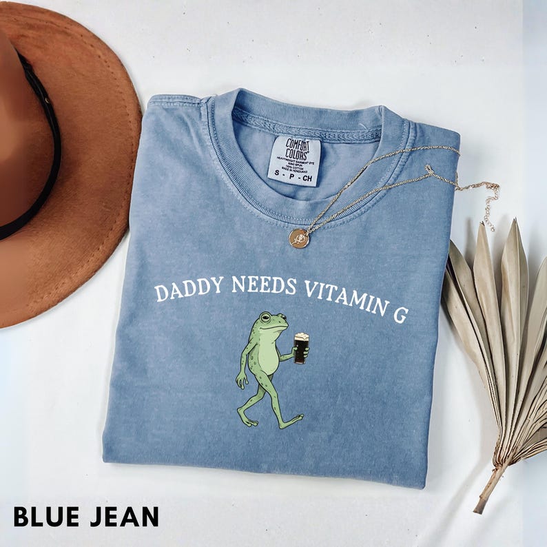 K&ouml;nnte beinhalten: Blaues Jeans Comfort Colors T-Shirt mit dem Text "DADDY NEEDS VITAMIN G" und einem Cartoon-Frosch, der ein dunkles Getr&auml;nk h&auml;lt. Eine goldene Halskette und ein brauner Hut sind ebenfalls sichtbar.