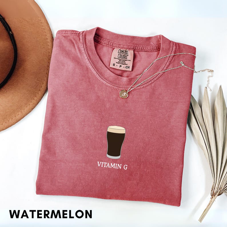 Pu&ograve; includere: T-shirt color anguria con girocollo e maniche corte. La maglietta presenta una grafica di una birra scura con la scritta "VITAMIN G" sotto. Sono visibili anche una collana d'argento e un cappello marrone.