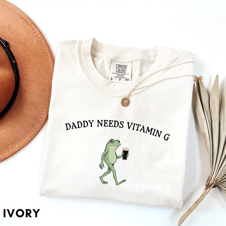 K&ouml;nnte beinhalten: Elfenbeinfarbenes Sweatshirt mit dem Text "DADDY NEEDS VITAMIN G" und einer Cartoon-Fr&ouml;schin, die ein dunkles Getr&auml;nk h&auml;lt. Das Shirt hat einen Rundhalsausschnitt und ist mit "COMFORT COLORS" gekennzeichnet. Eine goldene Halskette und ein brauner Hut sind ebenfalls zu sehen.