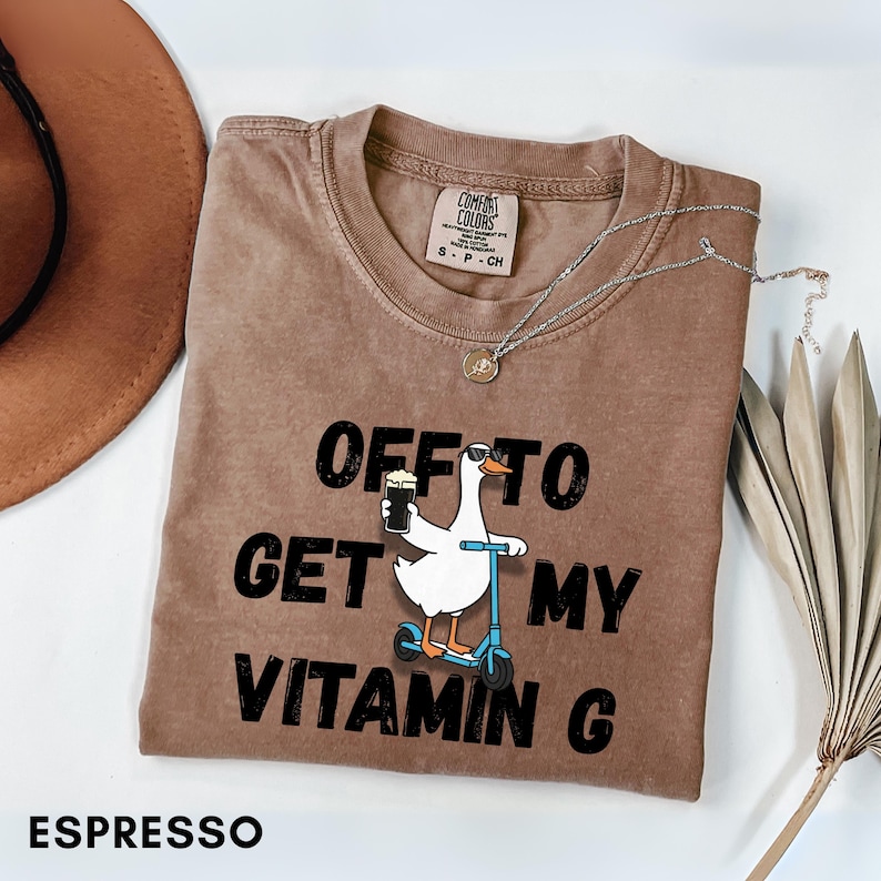 Puede incluir: Camiseta color espresso con la frase "OFF TO GET MY VITAMIN G" y un dibujo de un ganso con gafas de sol montado en un patinete, sosteniendo una cerveza. Tambi&eacute;n se ven un collar plateado y un sombrero marr&oacute;n.