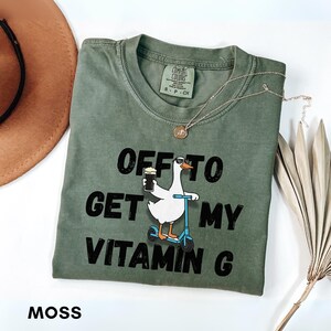 Puede incluir: Camiseta verde musgo con el texto "OFF TO GET MY VITAMIN G" y un dibujo de un ganso en un scooter con una cerveza. La camiseta tiene cuello redondo y mangas cortas. Una cadena de oro est&aacute; sobre la camiseta.