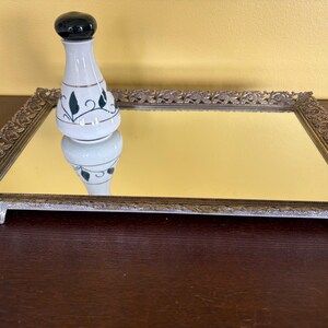 May include: A vintage mirrored vanity tray with an ornate silver-colored frame. A white and black ceramic perfume bottle with a green and gold leaf design sits on the tray. The tray is rectangular and rests on four small feet.