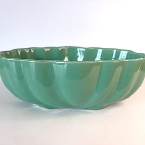 May include: A glossy, seafoam green ceramic bowl with a scalloped, fluted edge design. The bowl features a smooth, reflective finish and a rounded, wide shape with vertical indentations running along the exterior sides of the vessel.