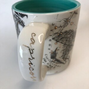 Puede incluir: Taza de cer&aacute;mica blanca con interior turquesa y asa. La taza presenta un dise&ntilde;o de zodiaco Capricornio negro y dorado con la palabra "Capricornio" en escritura dorada en el asa. La taza tiene una ilustraci&oacute;n detallada de la constelaci&oacute;n de Capricornio.