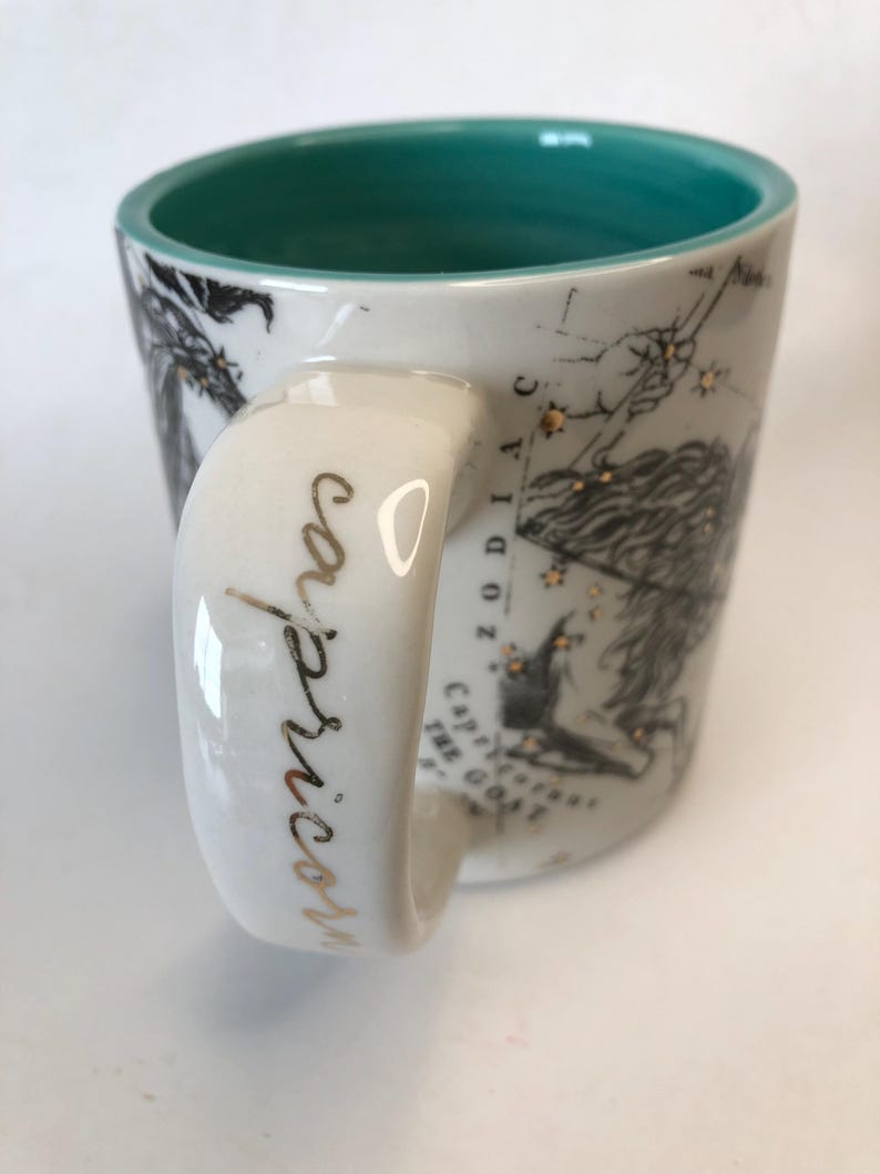 Puede incluir: Taza de cer&aacute;mica blanca con interior turquesa y asa. La taza presenta un dise&ntilde;o de zodiaco Capricornio negro y dorado con la palabra "Capricornio" en escritura dorada en el asa. La taza tiene una ilustraci&oacute;n detallada de la constelaci&oacute;n de Capricornio.
