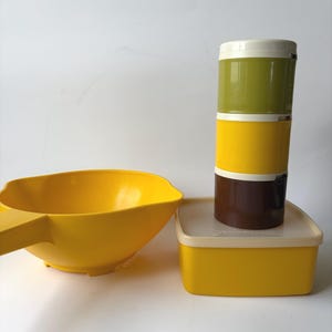 May include: A yellow plastic mixing bowl with a handle, a square yellow storage container with a cream lid, and a vertical stack of three cylindrical containers in olive green, yellow, and dark brown with a cream lid on top.