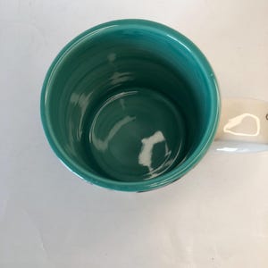 Puede incluir: Una taza de cer&aacute;mica turquesa con un asa blanca. El interior de la taza es de color turquesa liso. La taza est&aacute; sobre una superficie blanca.