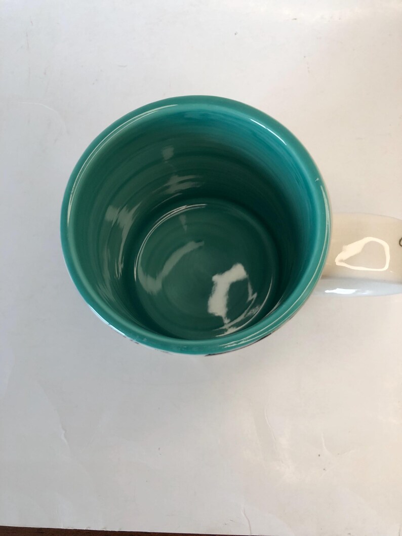 Puede incluir: Una taza de cer&aacute;mica turquesa con un asa blanca. El interior de la taza es de color turquesa liso. La taza est&aacute; sobre una superficie blanca.