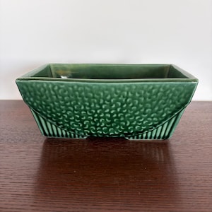 May include: A square, green ceramic planter with a textured, dotted pattern on the sides and a smooth, glossy finish. The planter has a geometric design with a slightly flared shape and is suitable for small plants or succulents.