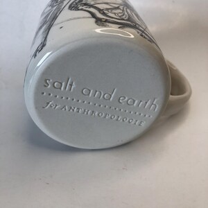 Puede incluir: Taza de cer&aacute;mica blanca con una ilustraci&oacute;n en blanco y negro. La base de la taza tiene el texto en relieve "salt and earth" y "for ANTHROPOLOGIE". La taza tiene un asa curva.