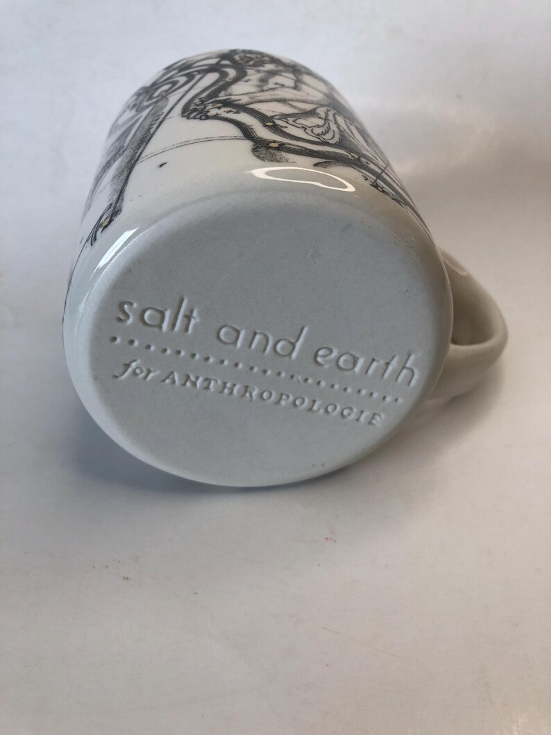 Puede incluir: Taza de cer&aacute;mica blanca con una ilustraci&oacute;n en blanco y negro. La base de la taza tiene el texto en relieve "salt and earth" y "for ANTHROPOLOGIE". La taza tiene un asa curva.