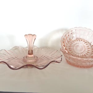 May include: A set of pink glass serving dishes. The set includes a tiered serving tray with a ruffled edge and a pedestal base, and two matching bowls with diamond-cut patterns.