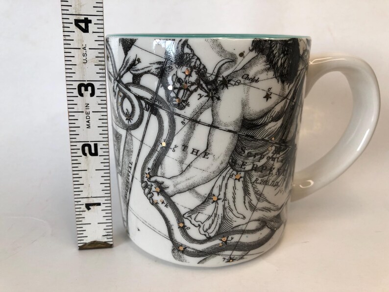 Puede incluir: Taza de cer&aacute;mica blanca con asa, con una ilustraci&oacute;n en blanco y negro de una figura mitol&oacute;gica con arco y flecha y constelaciones. La taza mide aproximadamente 7.6 cm de alto, con un borde turquesa.
