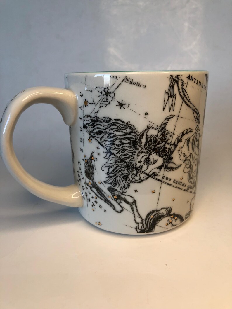 Puede incluir: Taza de cer&aacute;mica blanca con un dise&ntilde;o de constelaci&oacute;n del zodiaco en negro y dorado. La taza tiene un asa curva y un interior verde. El dise&ntilde;o incluye ilustraciones celestes y texto, con una est&eacute;tica vintage.