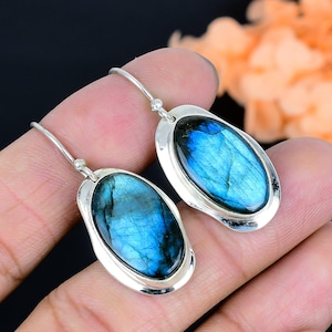 Puede incluir: Pendientes de plata con piedras de labradorita ovaladas. Las piedras muestran una vibrante iridiscencia azul, engastadas en un marco de plata pulida. Los pendientes tienen un diseño de gancho simple, mostrando la belleza natural de las piedras preciosas.
