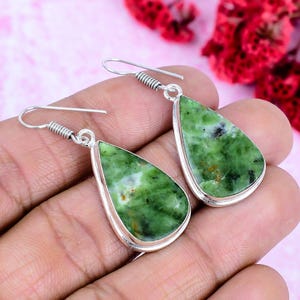 Puede incluir: Un par de pendientes en forma de lágrima con un marco de metal plateado. Los pendientes presentan una piedra verde con aspecto veteado. Los pendientes se muestran sobre un fondo rosa suave.