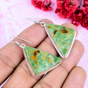 Peut inclure: Une paire de boucles d'oreilles argentées avec des pierres vertes triangulaires. Les pierres ont des inclusions marron et jaune et sont serties dans un cadre argenté. Les boucles d'oreilles sont présentées sur un fond rose.