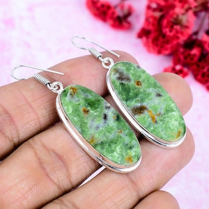 May include: A pair of oval-shaped earrings with a light green stone, accented with silver-colored metal. The stone has a speckled pattern with hints of brown and yellow. The earrings are displayed against a pink background.