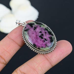 Op de afbeelding: Ovale hanger met een zilverkleurige beugel en een getextureerde rand. De hanger heeft een gepolijste edelsteen met een levendige mix van kleuren, waaronder roze, zwart en groen. De edelsteen is het middelpunt van de sieraden.
