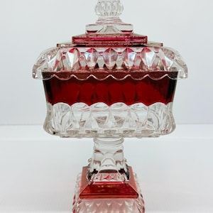 May include: A square glass candy dish with a lid, featuring a clear and deep red color scheme. The piece has a faceted, geometric pattern on the body, lid, and square base, supported by a central pedestal stem with a matching red and clear design.