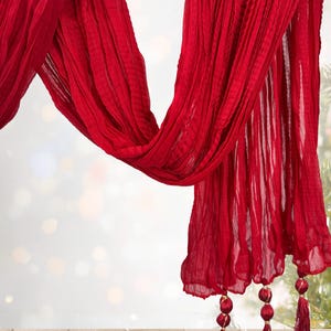 May include: A flowing, vibrant red scarf with a textured, pleated design. The scarf drapes gracefully, with decorative tassels at the ends. The fabric appears lightweight and airy, perfect for adding a pop of color.