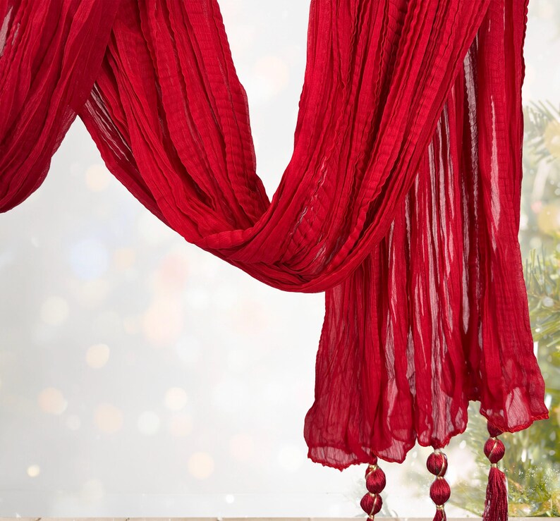 May include: A flowing, vibrant red scarf with a textured, pleated design. The scarf drapes gracefully, with decorative tassels at the ends. The fabric appears lightweight and airy, perfect for adding a pop of color.