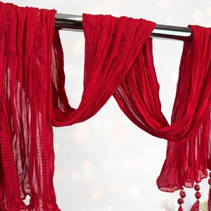 May include: A flowing, vibrant red scarf is draped over a silver rod. The fabric has a crinkled texture and hangs in graceful folds. Decorative tassels with red beads adorn the ends, adding a touch of elegance. The scarf is a striking accessory.