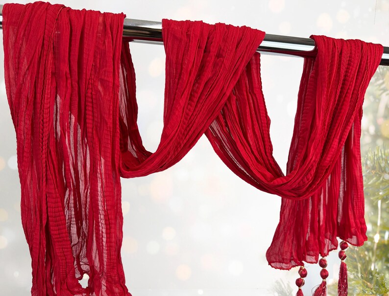 May include: A flowing, vibrant red scarf is draped over a silver rod. The fabric has a crinkled texture and hangs in graceful folds. Decorative tassels with red beads adorn the ends, adding a touch of elegance. The scarf is a striking accessory.