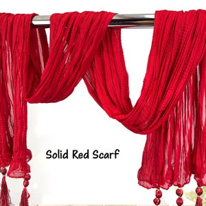 May include: A solid red scarf with a flowing, textured design. The scarf is draped over a metal bar, showcasing its length and fabric detail. The bottom edges feature decorative tassels and beaded accents. The text "Solid Red Scarf" is visible.