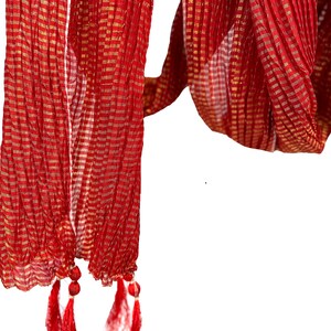 May include: A red scarf with a gold checkered pattern and red tassels. The scarf is made of a lightweight fabric and has a flowing design. The tassels are attached to the bottom edge of the scarf.