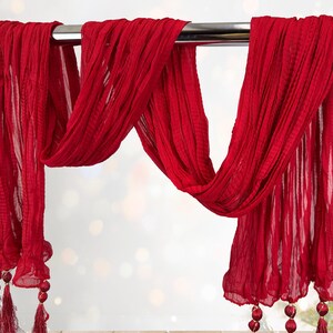May include: A flowing, red scarf with a textured, pleated design. The scarf is draped over a metal rod, with tassels and decorative beads at the ends. The fabric appears lightweight and airy, suitable for a decorative accent.