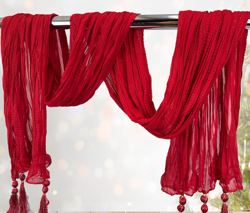 May include: A flowing, red scarf with a textured, pleated design. The scarf is draped over a metal rod, with tassels and decorative beads at the ends. The fabric appears lightweight and airy, suitable for a decorative accent.