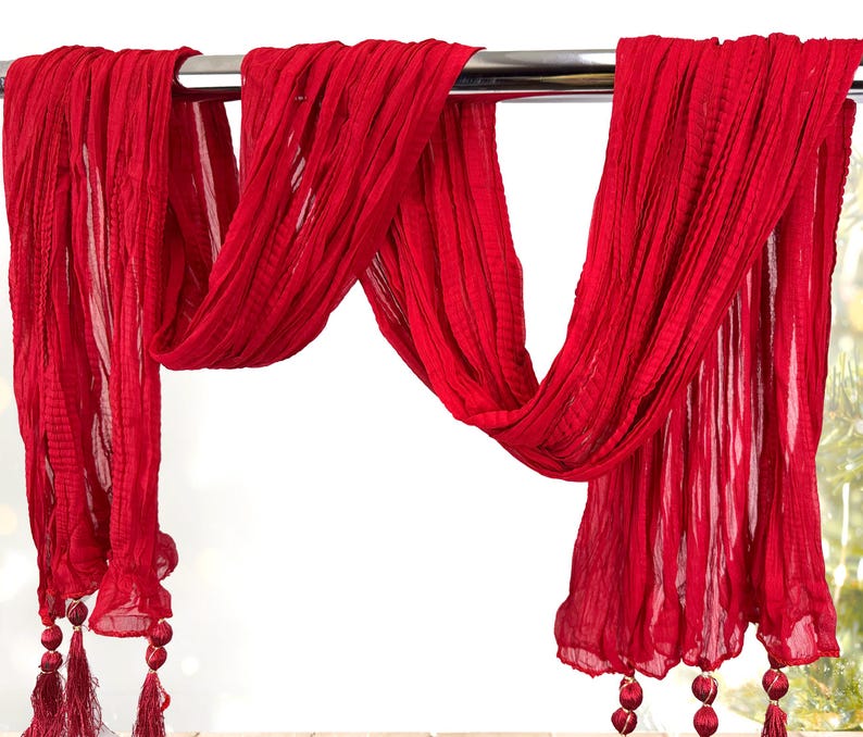 May include: A flowing, red scarf with a textured, pleated design. The scarf is draped over a metal bar, with tassels and decorative beads at the ends. The fabric appears lightweight and airy, suitable for a fashion accessory.