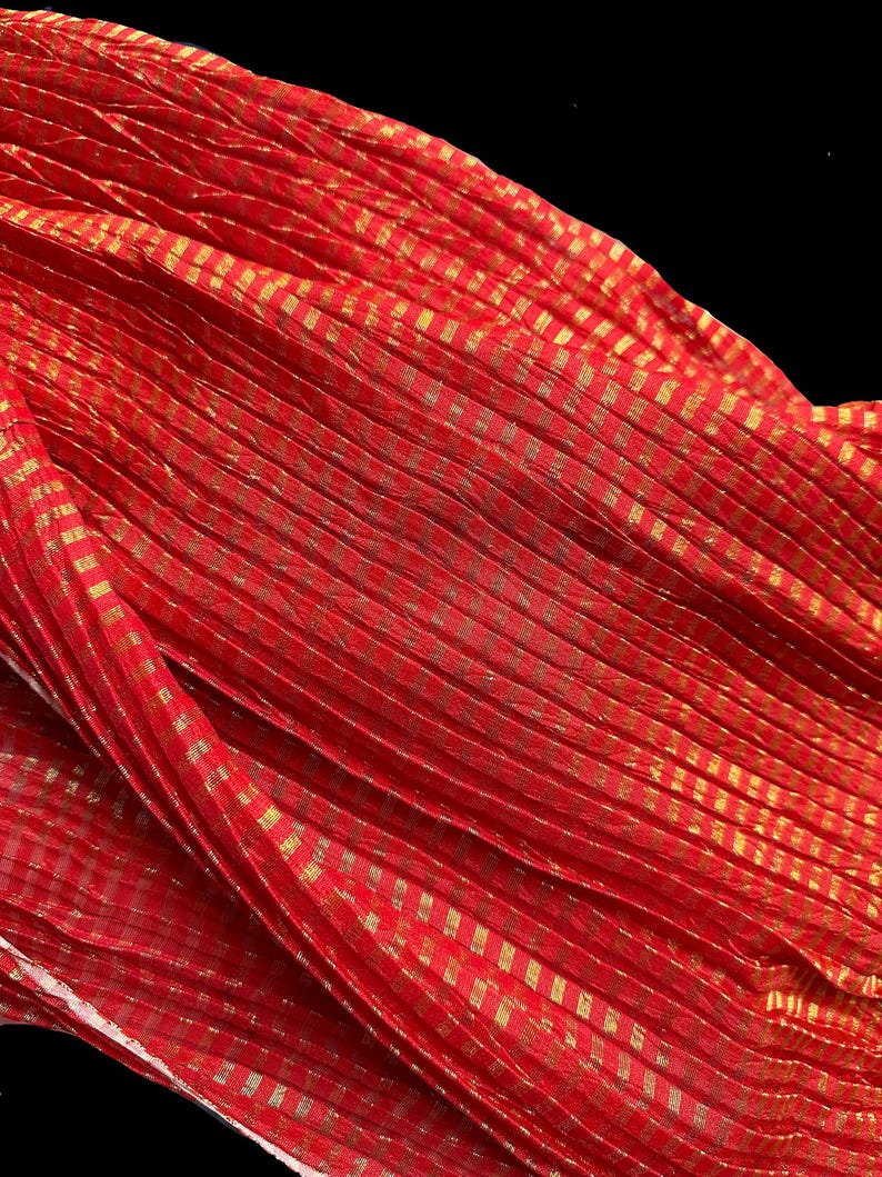 May include: A vibrant red fabric with a gold checkered pattern. The fabric is draped, creating folds and shadows that highlight the texture and design. The gold accents add a touch of elegance to the overall look.