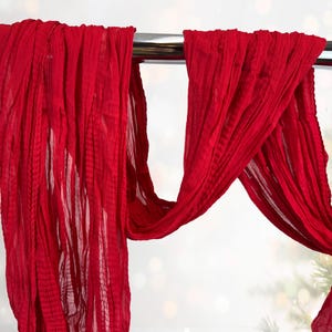 May include: Bright red, flowing fabric draped over a silver bar. The fabric has a textured, pleated appearance, creating a sense of movement. The background is blurred with soft, out-of-focus lights and hints of greenery.