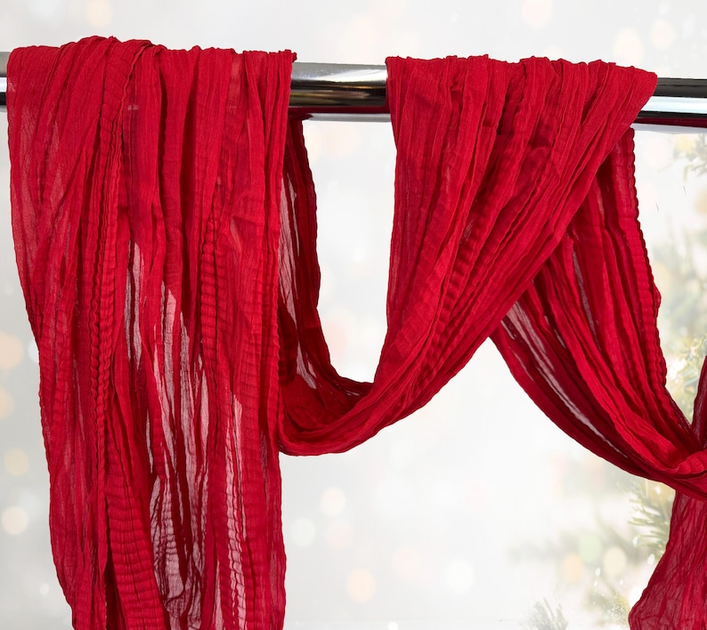 May include: Bright red, flowing fabric draped over a silver bar. The fabric has a textured, pleated appearance, creating a sense of movement. The background is blurred with soft, out-of-focus lights and hints of greenery.