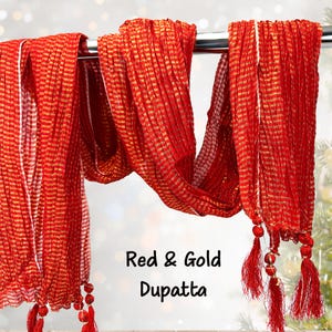 May include: A red and gold dupatta, a traditional Indian scarf, hangs from a metal rod. The scarf features a striped pattern with gold accents and red tassels at the ends. The text "Red & Gold Dupatta" is visible.