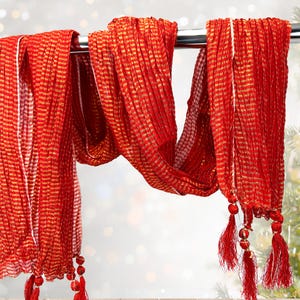May include: A vibrant red scarf with gold stripes hangs from a metal rod. The scarf has a textured, pleated design and is adorned with red tassels at the ends. The background is blurred, suggesting a soft focus.