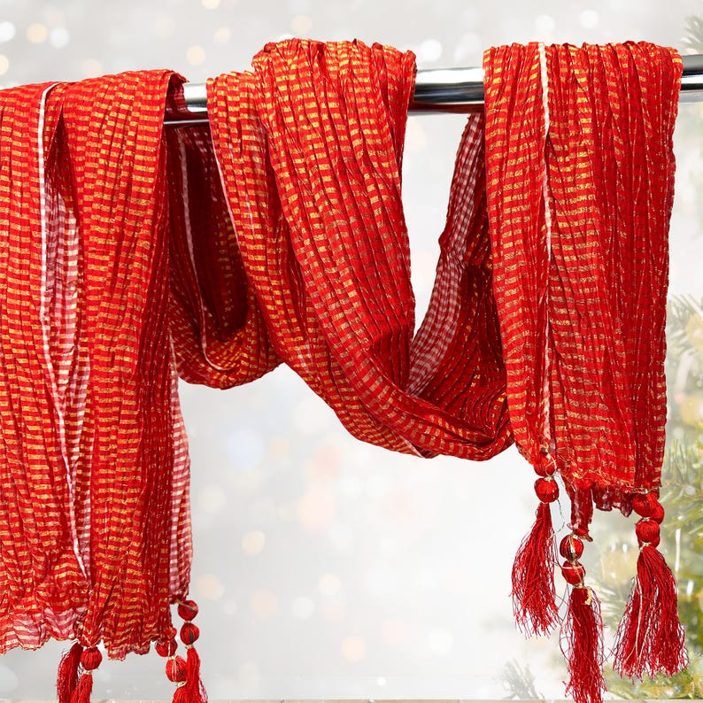 May include: A vibrant red scarf with gold stripes hangs from a metal rod. The scarf has a textured, pleated design and is adorned with red tassels at the ends. The background is blurred, suggesting a soft focus.