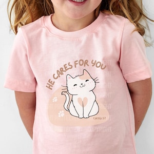 Könnte beinhalten: Ein rosa T-Shirt mit einer weißen Katzenillustration und dem Text "He cares for you".