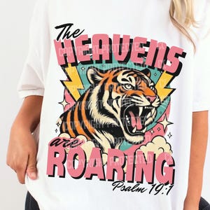 Pode incluir: Camiseta branca com um gráfico de um tigre rugindo e o texto "The Heavens are Roaring" em rosa e preto. O design inclui raios e o versículo bíblico Salmo 19:1.