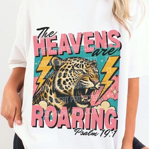 Könnte beinhalten: Weißes T-Shirt mit einer Grafik eines brüllenden Leoparden und dem Text "The Heavens are Roaring" in Rosa und Schwarz. Das Design beinhaltet Blitze und den Bibelvers Psalm 19:1.