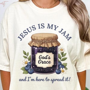 May include: Cream-colored t-shirt with the text "JESUS IS MY JAM" above a graphic of a jam jar labeled "God's Grace." The design includes blueberries and flowers. The phrase "and I'm here to spread it!" is printed below the jar.
