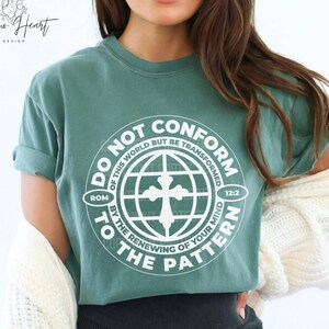 Puede incluir: Una camiseta verde salvia con el texto blanco "DO NOT CONFORM TO THE PATTERN" en un diseño circular. El diseño incluye una cruz y las palabras "ROM 12:2". La camiseta es de estilo casual.