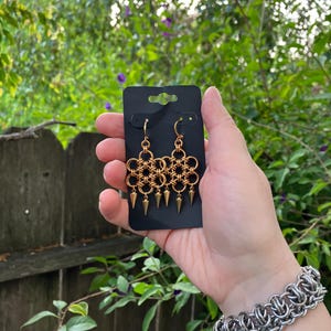 May include: Gold-colored chainmail earrings with a floral design and dangling cone-shaped accents. The earrings are displayed on a black card, with a silver chainmail bracelet visible in the background. The earrings are hanging in front of a green leafy background.