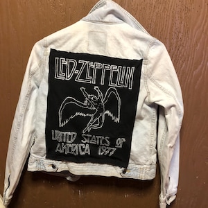 May include: Light blue denim jacket with a black patch featuring the Led Zeppelin logo and a winged figure. The patch also includes the text "UNITED STATES OF AMERICA 1977". The jacket is hanging on a white hanger.